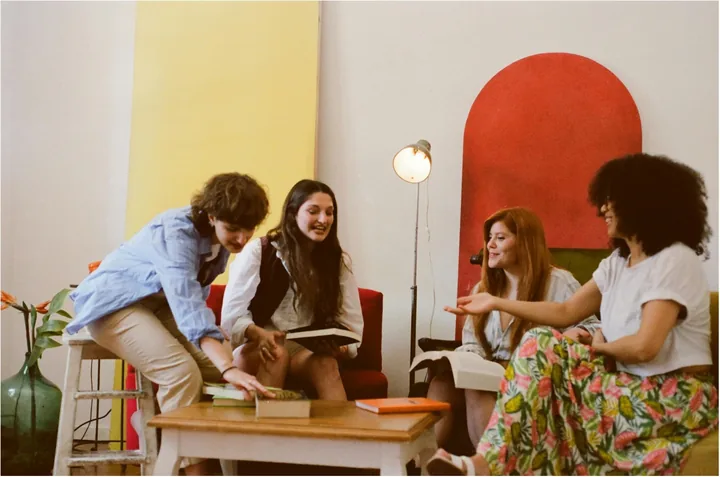 People chatting around a table with books, against yellow and red walls.
