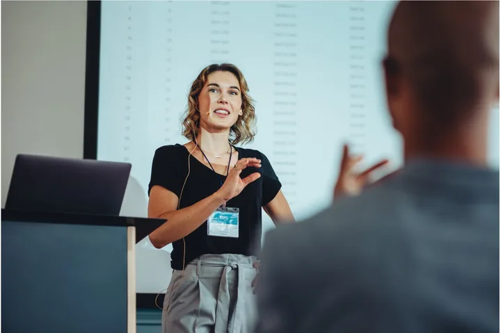 A woman speaking to an audience with a headset microphone.